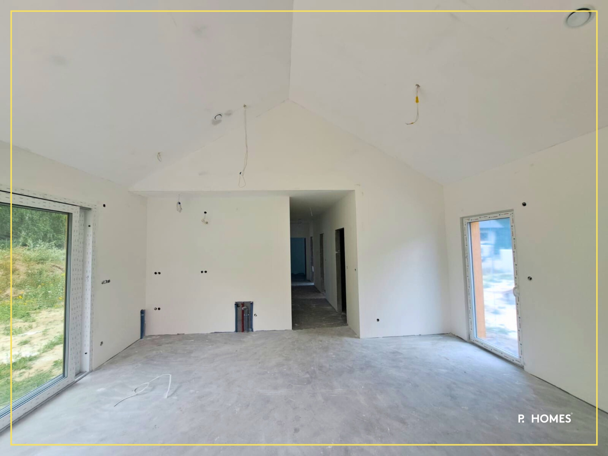 Wnętrze domu w standardzie deweloperskim: surowe ściany z instalacjami, betonowa podłoga, okna PVC. Widok na kolejne pomieszczenia w głębi. Oświetlenie sufitowe.