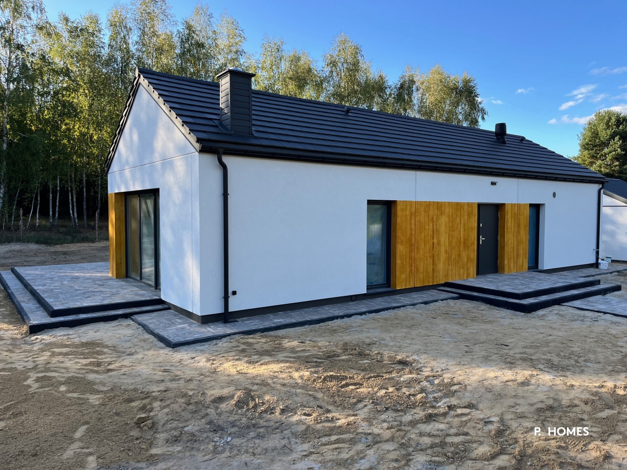 Nowoczesny, parterowy dom z białymi ścianami, czarnym dachem i drewnianymi akcentami, otoczony zielenią. Widoczne tarasy z kostki brukowej i fragment ogrodu.