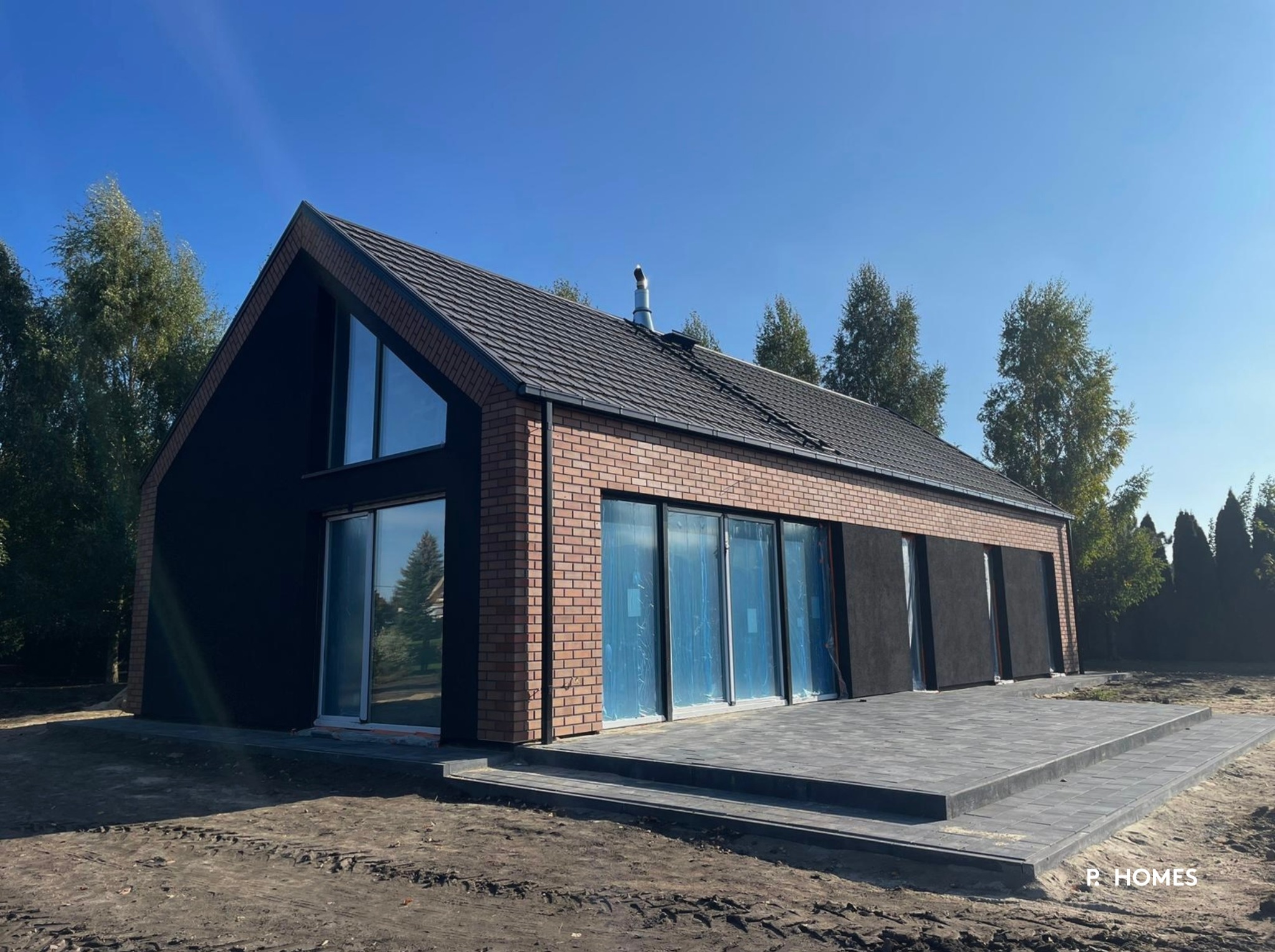Nowoczesny dom jednorodzinny z czarną elewacją i ceglanym wykończeniem, duże przeszklenia, taras z kostki brukowej, projekt P.Homes w słoneczny dzień.
