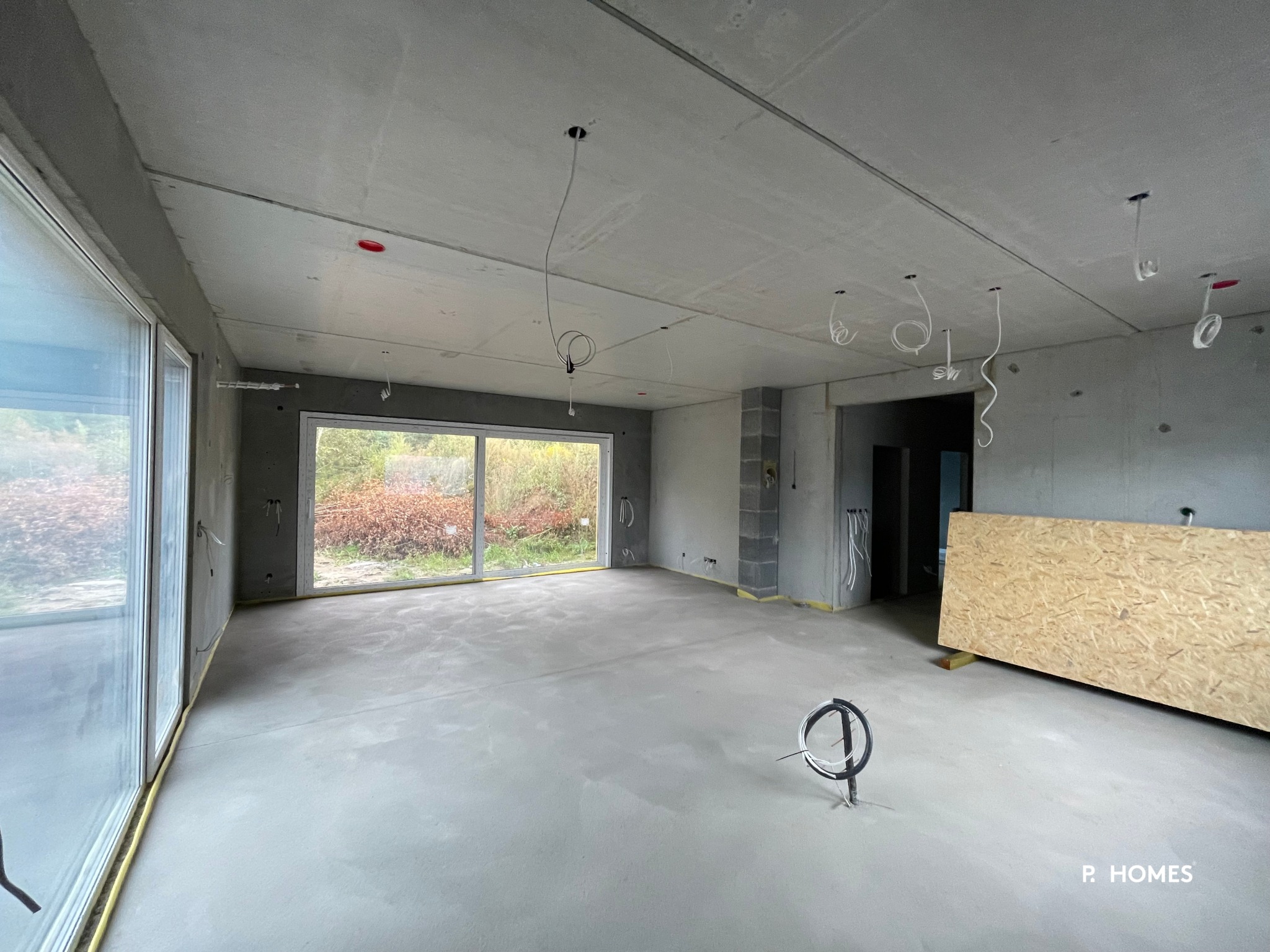 Surowe wnętrze domu w stanie deweloperskim z betonowymi ścianami, podłogą i dużymi oknami. Widoczne wystające przewody elektryczne i płyta OSB oparta o ścianę.