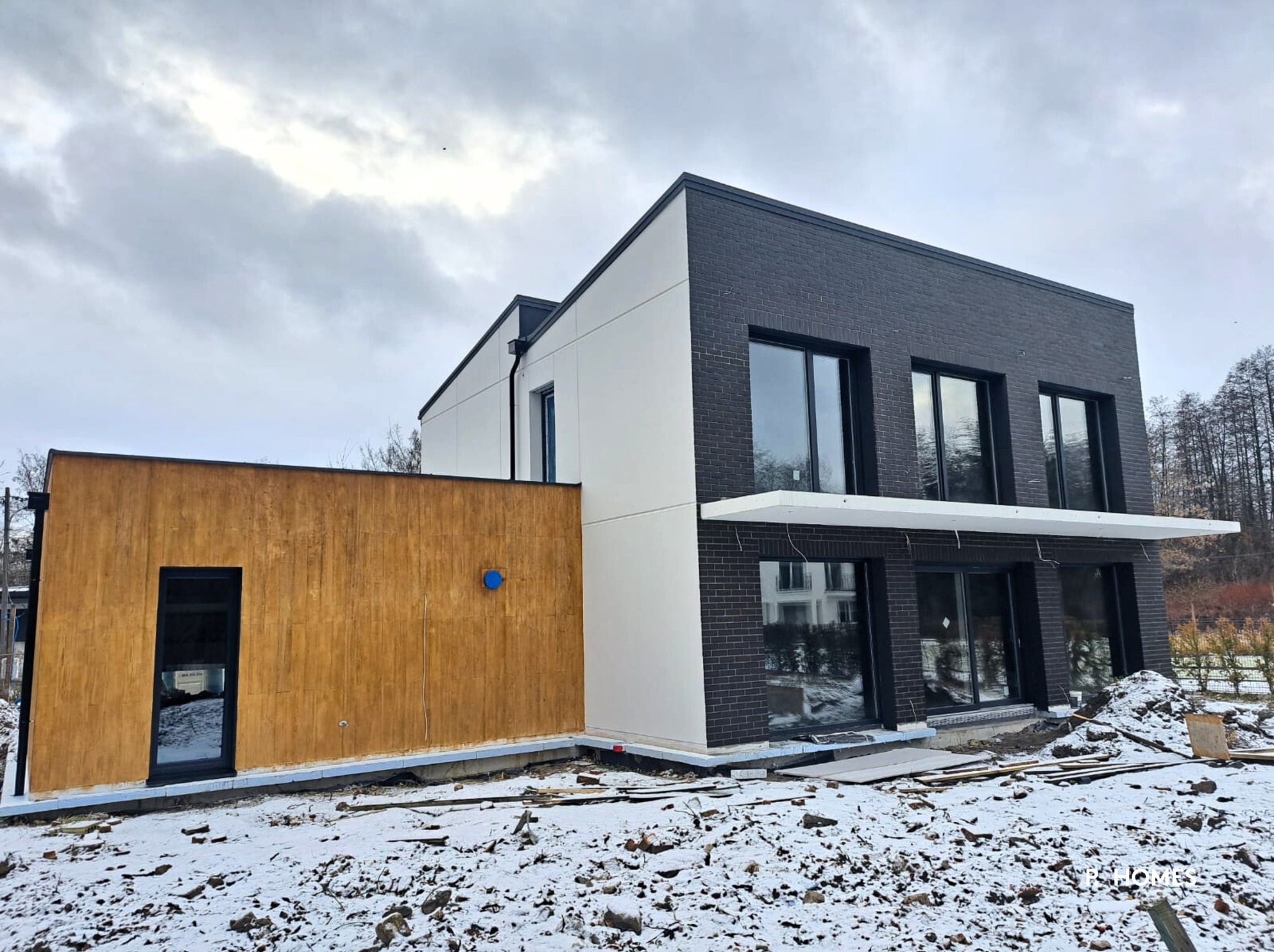 Nowoczesny dom z elementami drewna, czarnej cegły i bieli, częściowo zasypany śniegiem. Widoczne okna i drzwi w czarnych ramach, surowy krajobraz.