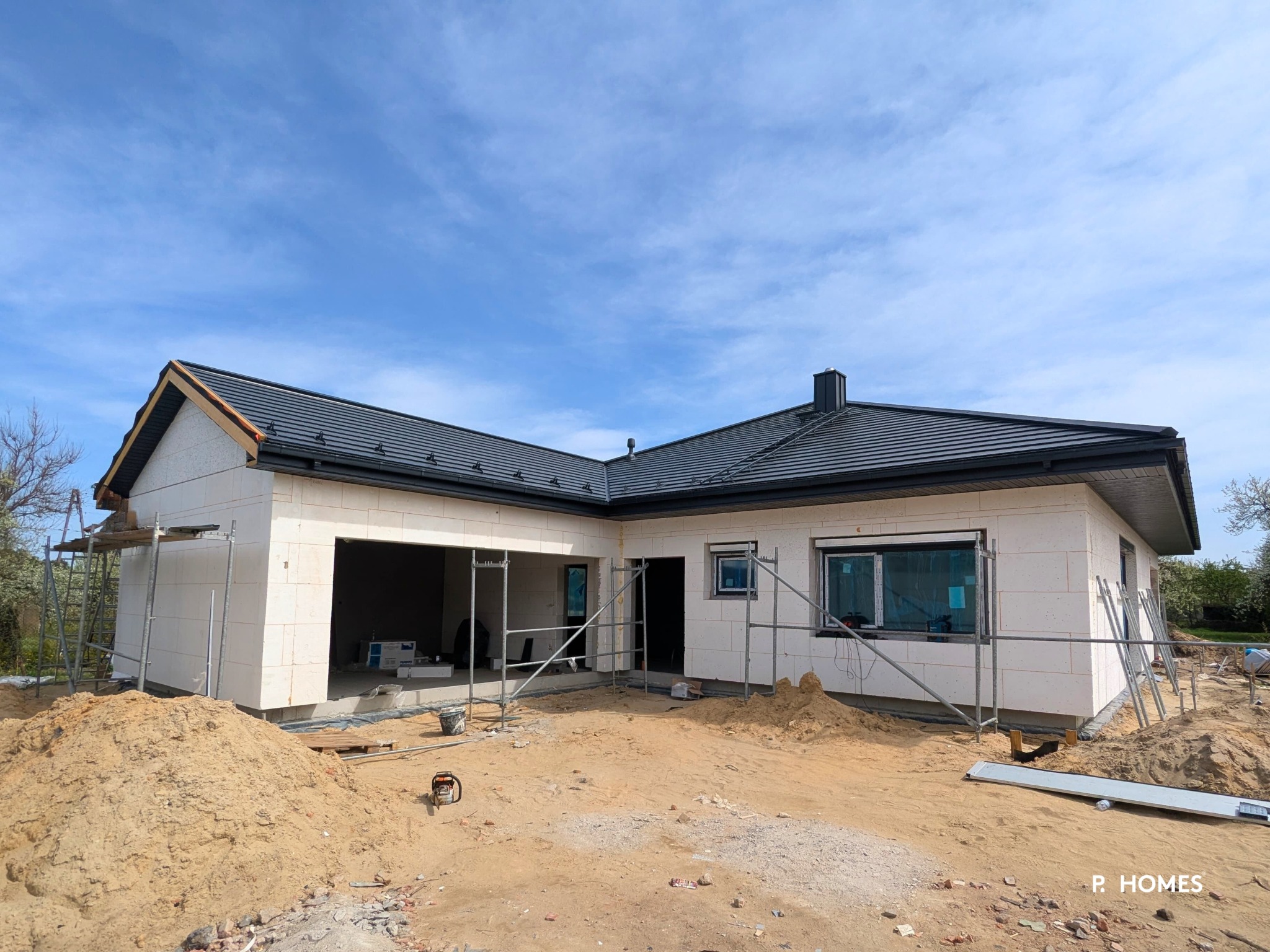 Niedokończony dom parterowy z białych bloczków, z ciemną dachówką, widoczne rusztowania i piasek. Częściowo zamontowane okna z folią ochronną.