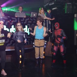 Dj Monde - Impreza kostiumowa: grupa osób przebranych za postacie z gier i filmów, w tle DJ-ka z oświetleniem. Uczestnicy w kostiumach DeadPool, Lary Croft i innych postaci.