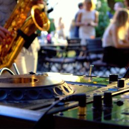 Dj Monde - Dj miksuje muzykę na imprezie plenerowej, w tle saksofonista i goście. Scena oświetlona, sprzęt nagłośnieniowy na pierwszym planie.