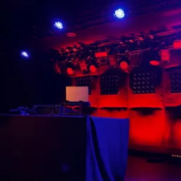 Scena oświetlona czerwienią i błękitem, z laptopem Dell na stole DJ-skim. Widoczne reflektory i elementy nagłośnienia. Scena gotowa na występ.