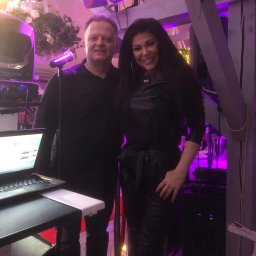 Dj Monde - Duet muzyczny na scenie z laptopem, mikrofonem i oświetleniem w tle. Kobieta w czarnym stroju, mężczyzna w czarnej koszuli. Scena oświetlona fioletowym światłem.