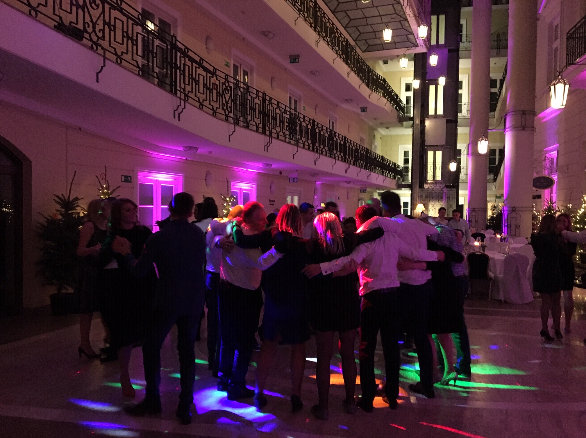 Rozświetlona sala balowa w Warszawie z bawiącymi się ludźmi, oświetlenie sceniczne i dekoracje świąteczne tworzą radosny nastrój.