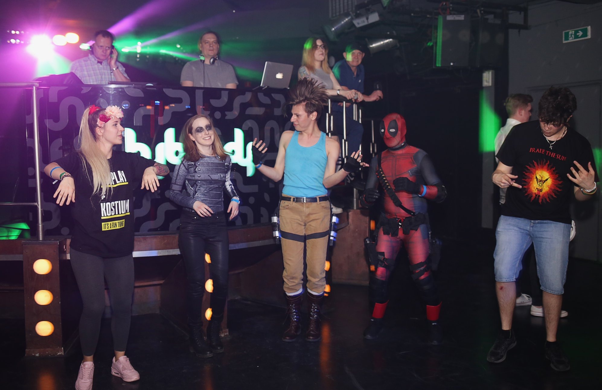 Impreza kostiumowa: grupa osób przebranych za postacie z gier i filmów, w tle DJ-ka z oświetleniem. Uczestnicy w kostiumach DeadPool, Lary Croft i innych postaci.