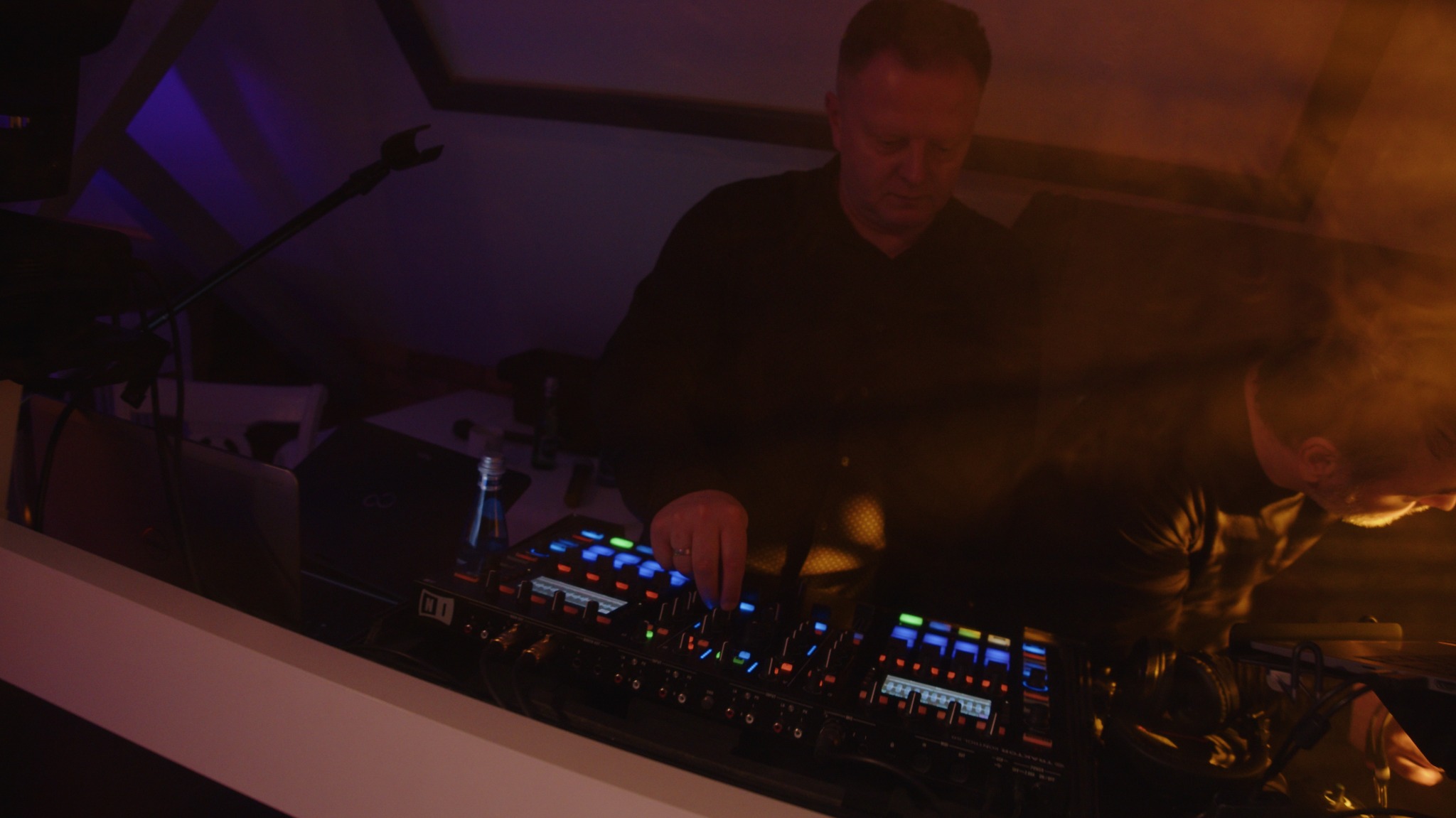 DJ miksuje muzykę na profesjonalnym sprzęcie w klubowym oświetleniu. Widoczny mikrofon i butelka wody. Ujęcie z góry, skupione na konsoli.