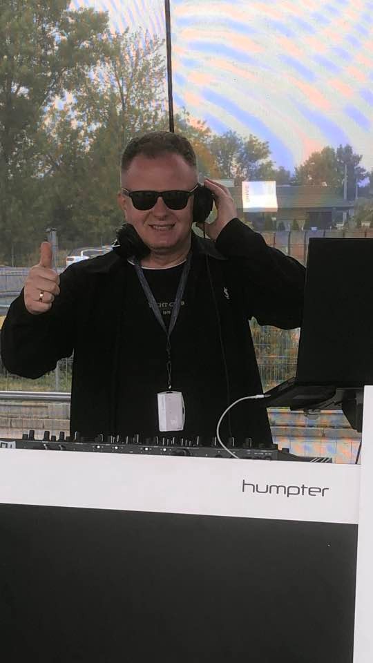 Uśmiechnięty DJ w czarnych okularach i słuchawkach, z uniesionym kciukiem, za konsoletą marki 'humper' podczas imprezy plenerowej.