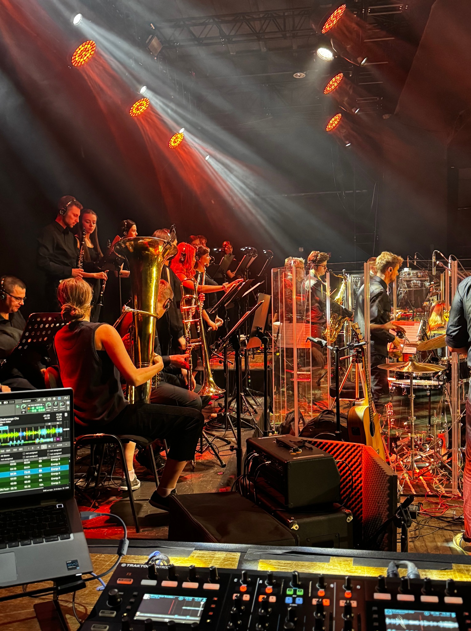 Orkiestra symfoniczna na scenie w czerwonym oświetleniu. Widoczny laptop z oprogramowaniem muzycznym i konsoleta mikserska.