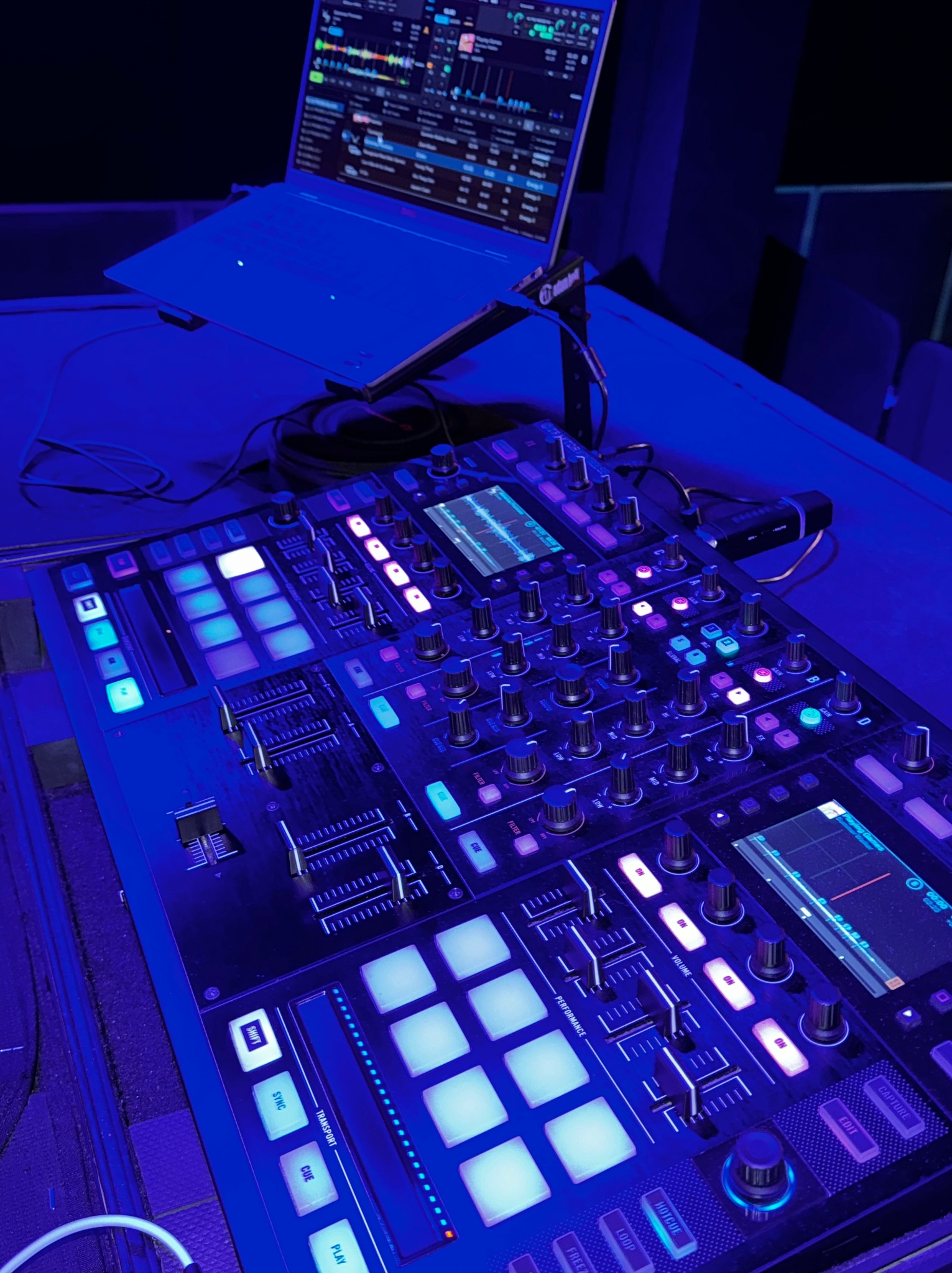 Sprzęt DJ-ski oświetlony na niebiesko: mikser, laptop z oprogramowaniem, kable. Widok z góry na stanowisko pracy DJ-a w klubie lub studio.