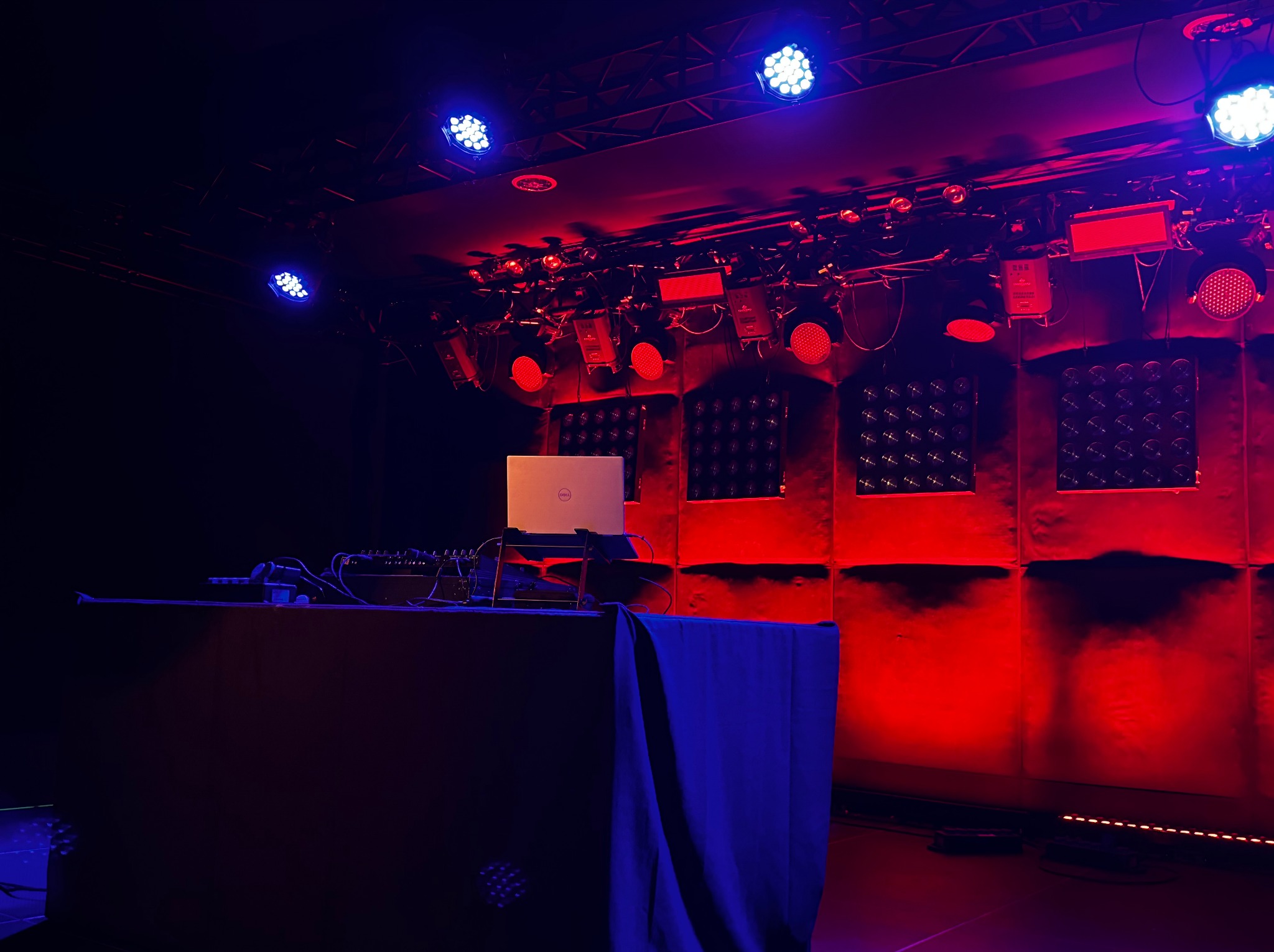 Scena oświetlona czerwienią i błękitem, z laptopem Dell na stole DJ-skim. Widoczne reflektory i elementy nagłośnienia. Scena gotowa na występ.