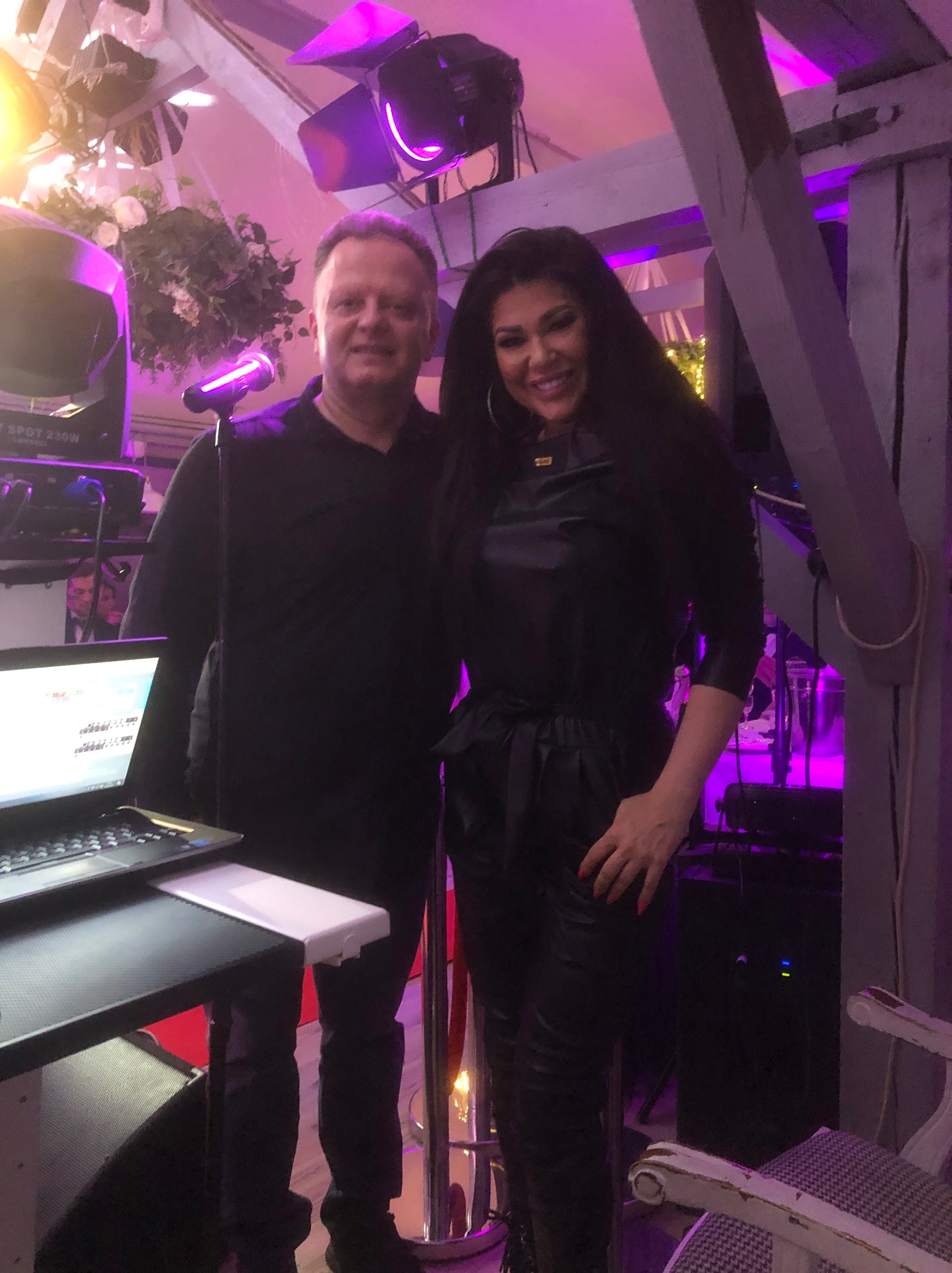 Duet muzyczny na scenie z laptopem, mikrofonem i oświetleniem w tle. Kobieta w czarnym stroju, mężczyzna w czarnej koszuli. Scena oświetlona fioletowym światłem.