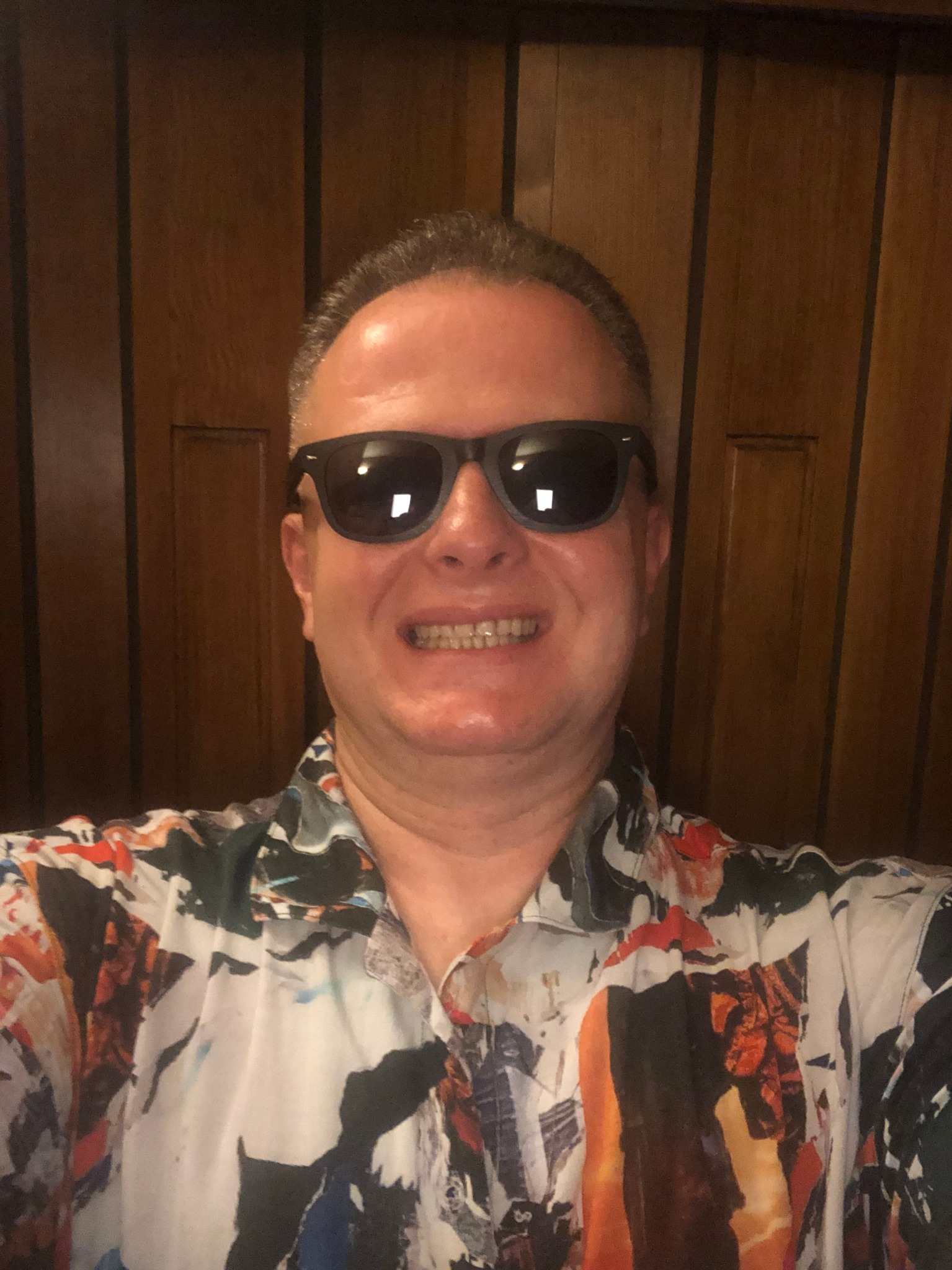 Uśmiechnięty mężczyzna w okularach przeciwsłonecznych i wzorzystej koszuli, na tle drewnianej ściany. Widoczne zęby i fragmenty koszuli z abstrakcyjnym deseniem.