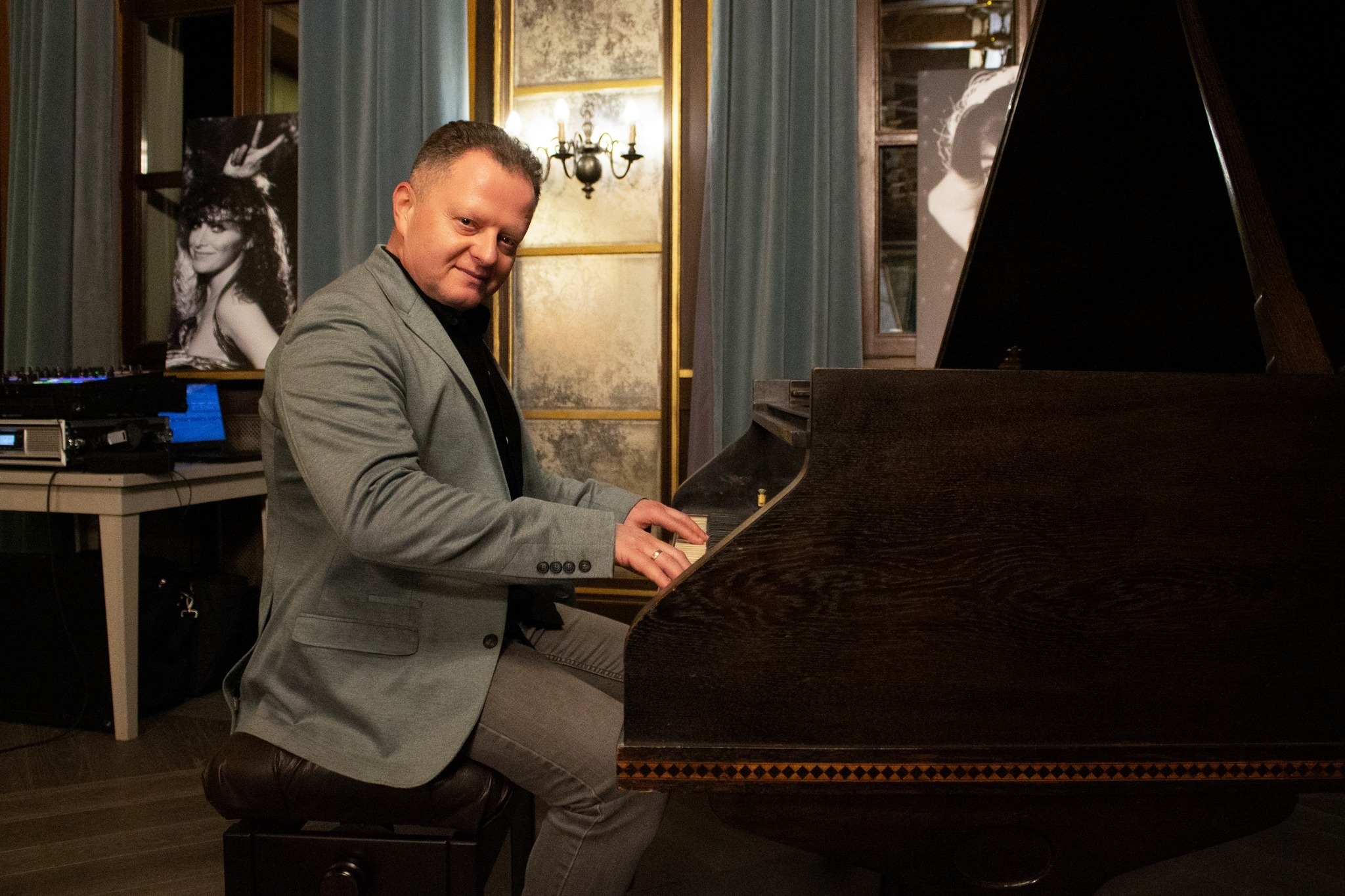 Pianista w szarym garniturze gra na fortepianie w eleganckim wnętrzu z dekoracyjnymi zasłonami i portretami w tle, tworząc nastrojową atmosferę.