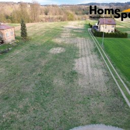 HomeSpace Nieruchomości - Szeroki kadr z lotu ptaka ukazuje dwa domy jednorodzinne na rozległym terenie zielonym. W tle las i pagórki. Logo firmy w prawym górnym rogu.