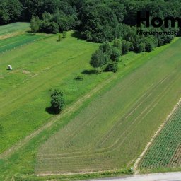 HomeSpace Nieruchomości - Zdjęcie z drona: budowa domu w stanie surowym otoczona zielonymi polami, lasem i logo agencji nieruchomości w prawym górnym rogu.