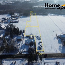 HomeSpace Nieruchomości - Zimowy widok z lotu ptaka na działkę z naniesionymi wymiarami (34m, 50m, 17m) w żółtych liniach. W tle zabudowa jednorodzinna i logo Home Space Nieruchomości.