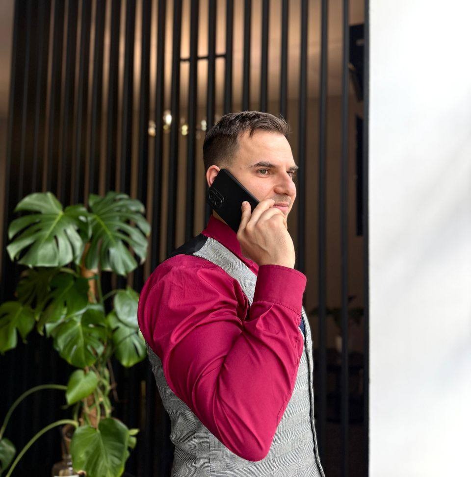 Mężczyzna w bordowej koszuli i kraciastej kamizelce rozmawia przez telefon na tle nowoczesnego wnętrza z rośliną monstera.