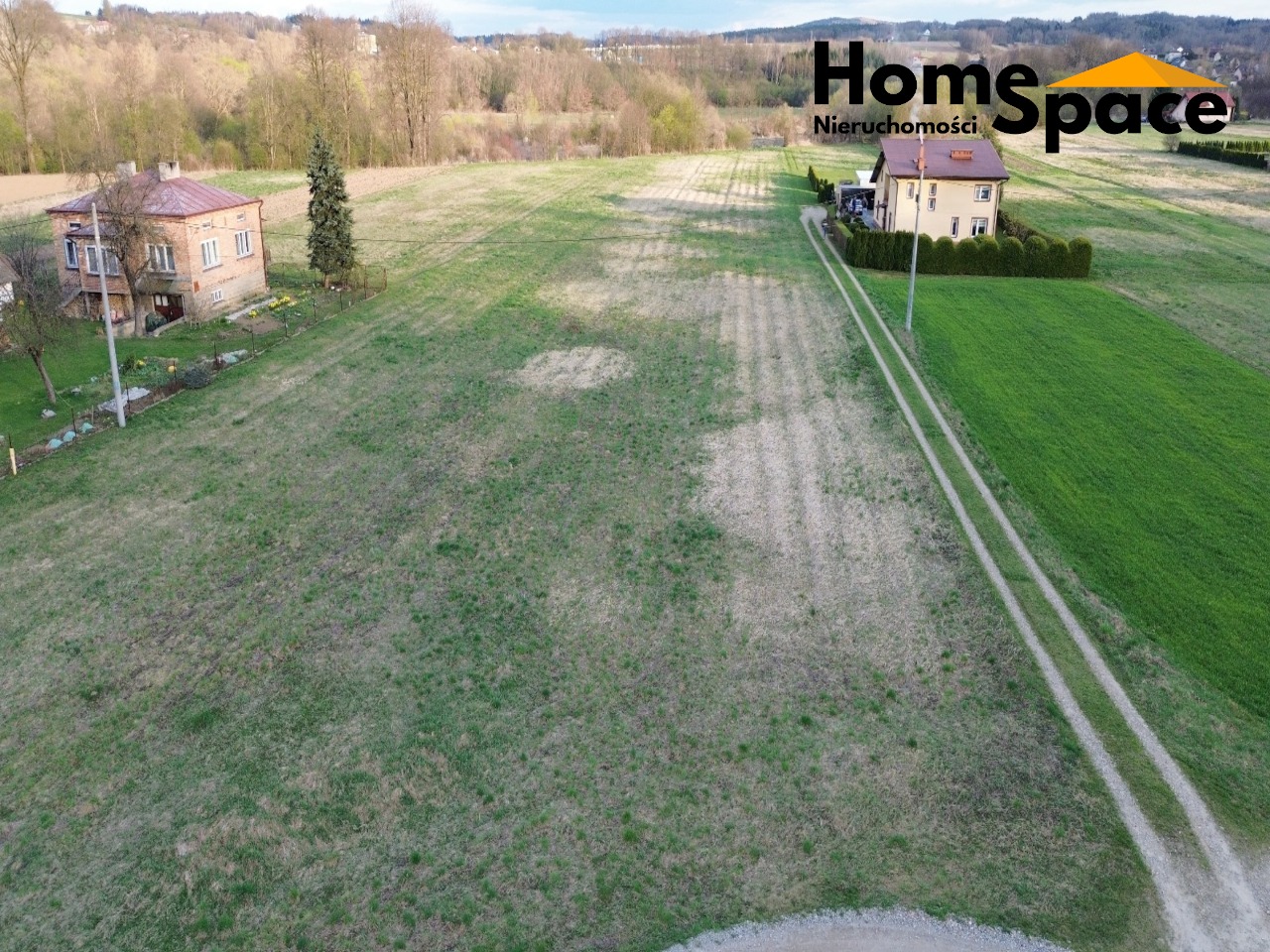 Szeroki kadr z lotu ptaka ukazuje dwa domy jednorodzinne na rozległym terenie zielonym. W tle las i pagórki. Logo firmy w prawym górnym rogu.