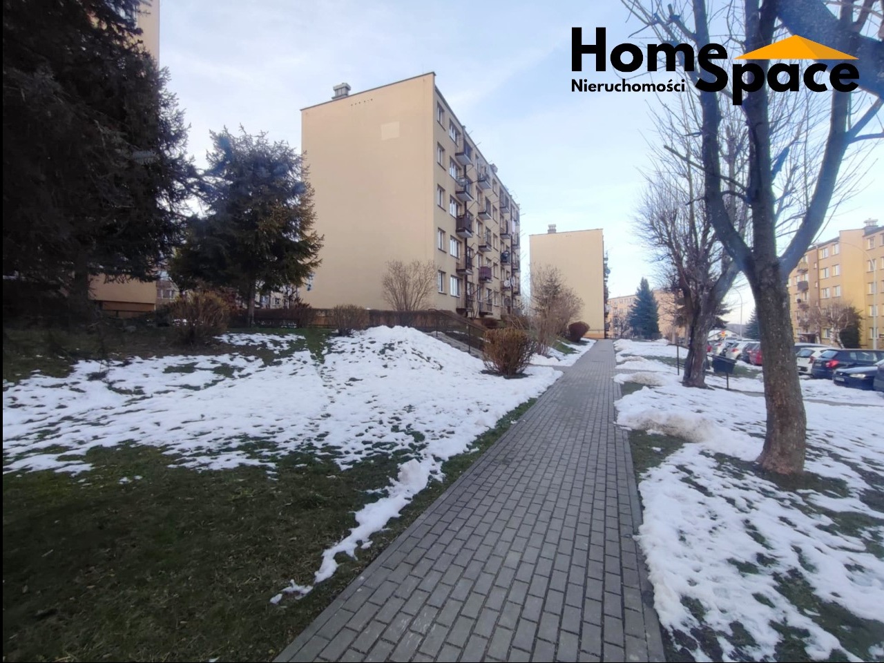 Blok mieszkalny z logo Home Space w zimowej scenerii. Kostka brukowa prowadzi wzdłuż ośnieżonego trawnika. Częściowo odsłonięte drzewa i zaparkowane auta w tle.