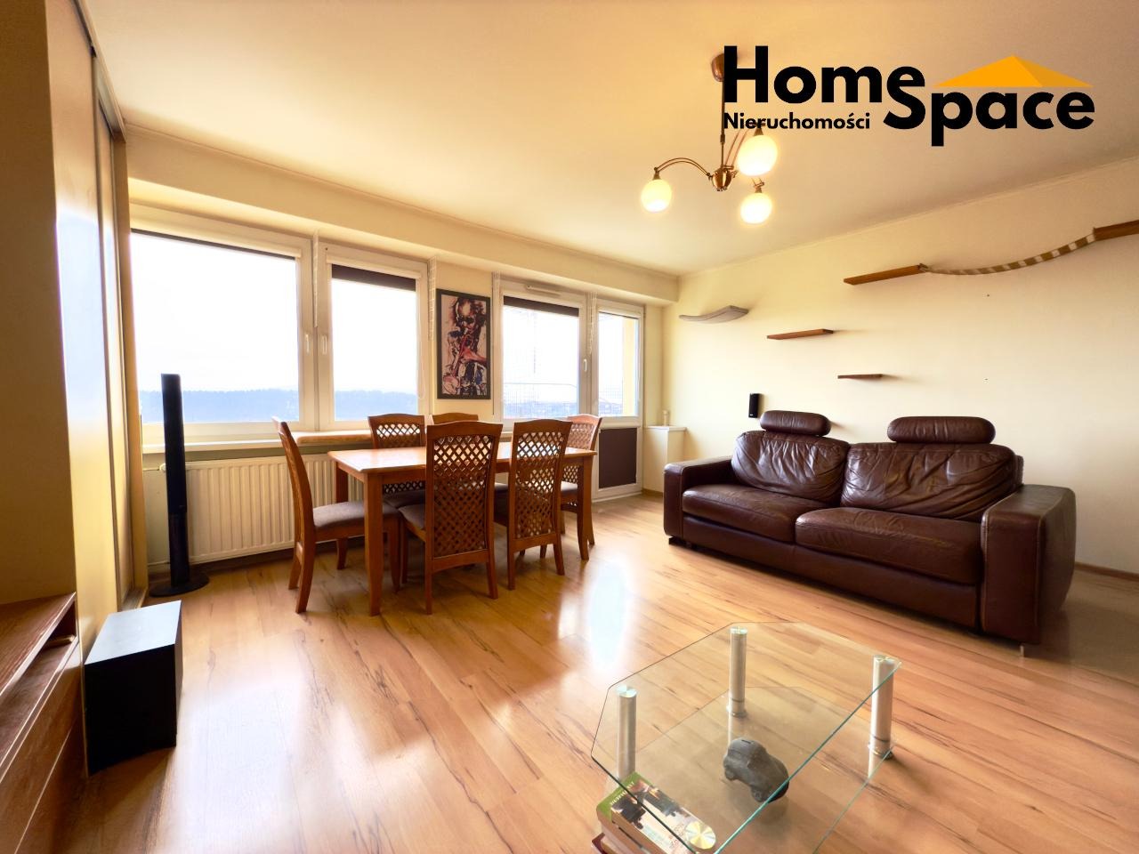 Jasne wnętrze z drewnianym stołem, skórzaną kanapą i panoramicznymi oknami. Na ścianie półki dla kota. Logo firmy Home Space Nieruchomości w prawym górnym rogu.
