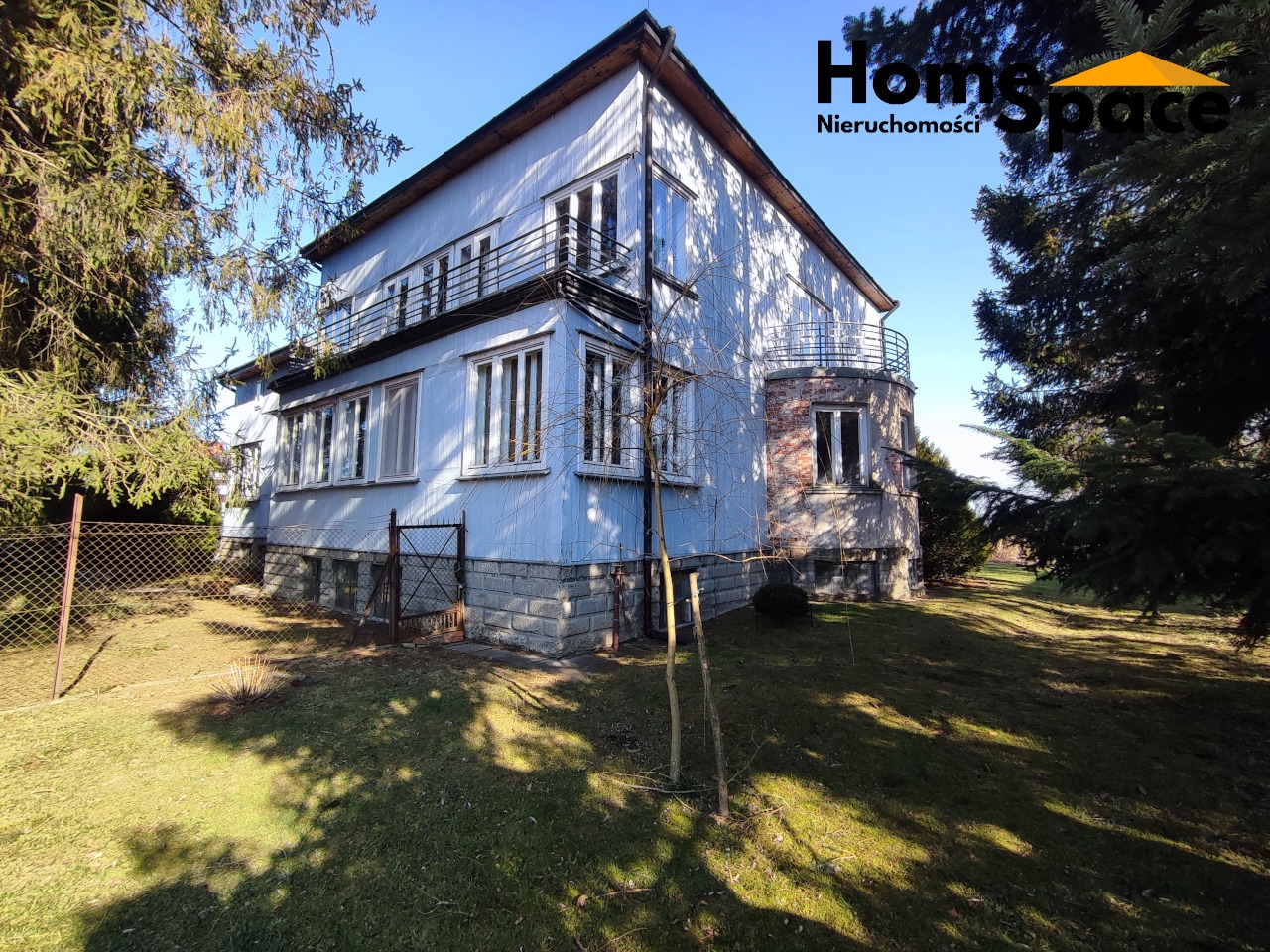 Dwupiętrowy dom z balkonami w Krygu, Małopolska. Budynek z jasną elewacją, częściowo obłożony kamieniem, otoczony zielenią i drzewami. Widok z perspektywy ogrodu.