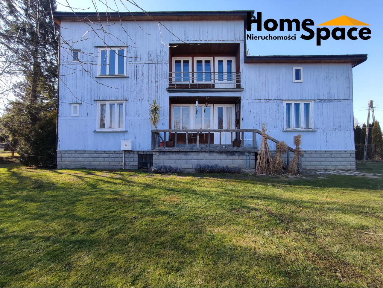 Dwupiętrowy dom z balkonami i logo 'Home Space Nieruchomości' w prawym górnym rogu, otoczony zielonym trawnikiem w słoneczny dzień.
