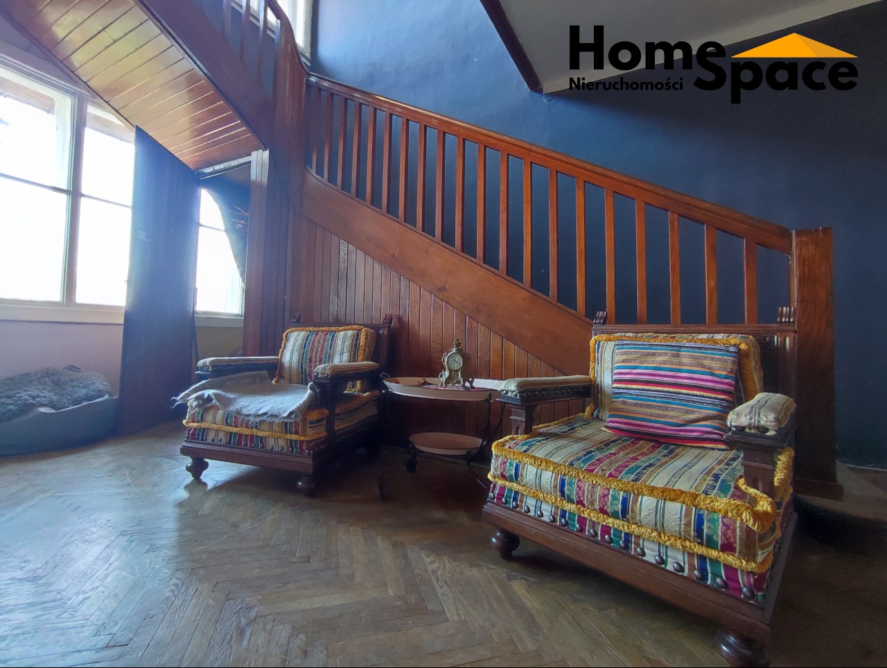 Rustykalne wnętrze z drewnianymi schodami, dwoma fotelami w stylu vintage i parkietem w jodełkę. W tle logo HomeSpace Nieruchomości.