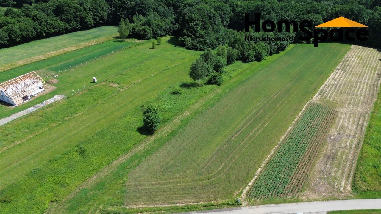 Zdjęcie z drona: budowa domu w stanie surowym otoczona zielonymi polami, lasem i logo agencji nieruchomości w prawym górnym rogu.
