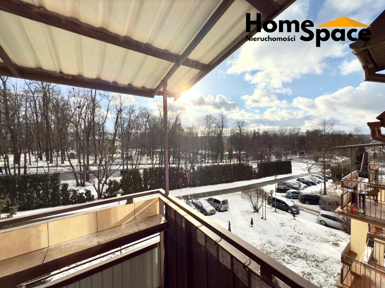 Zimowy widok z balkonu na osiedle z parkingiem i lasem w tle. Słońce przebija przez chmury, oświetlając śnieżny krajobraz. Logo firmy Home Space w prawym górnym rogu.