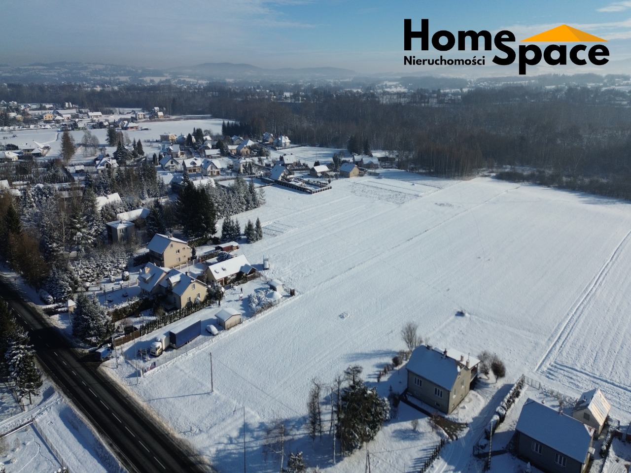 Zimowy krajobraz z lotu ptaka: ośnieżone domy, pola i lasy w oddali. Logo Home Space Nieruchomości w prawym górnym rogu.