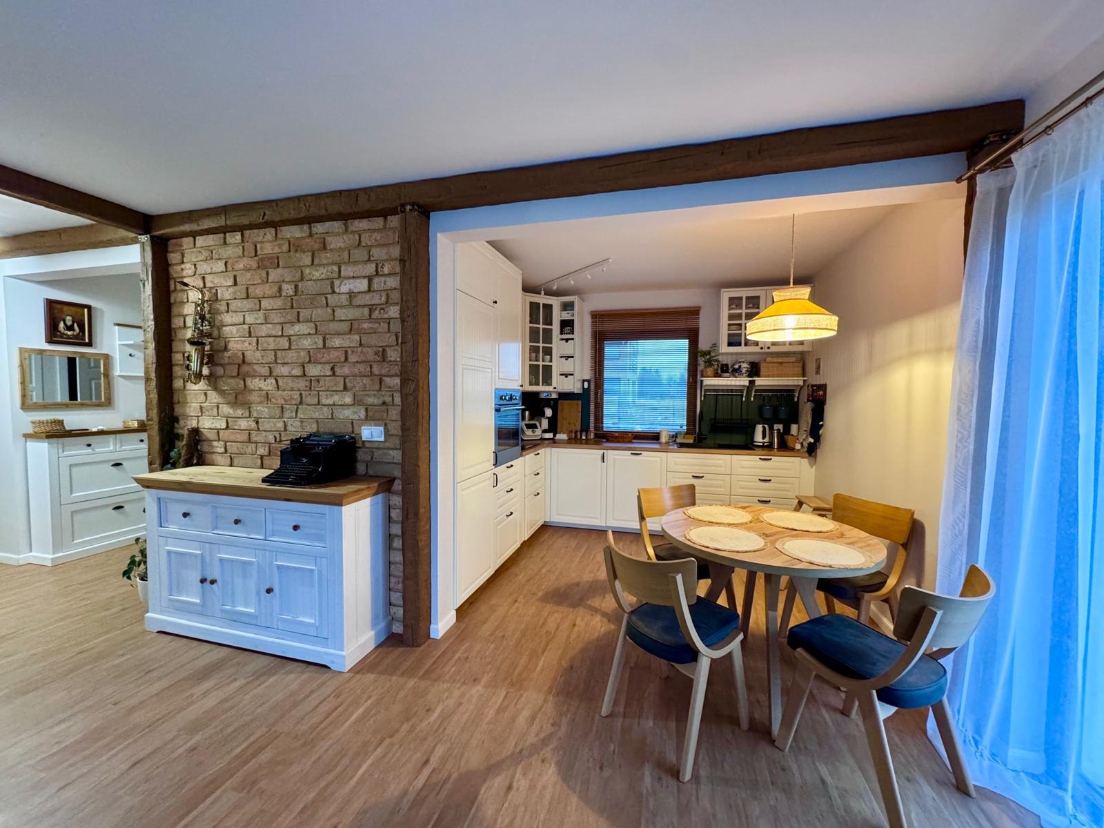 Jasna kuchnia z drewnianymi belkami i ceglaną ścianą. Stół z krzesłami, białe szafki i sprzęty. Na komodzie stoi czarna maszyna do pisania. Widok na salon.