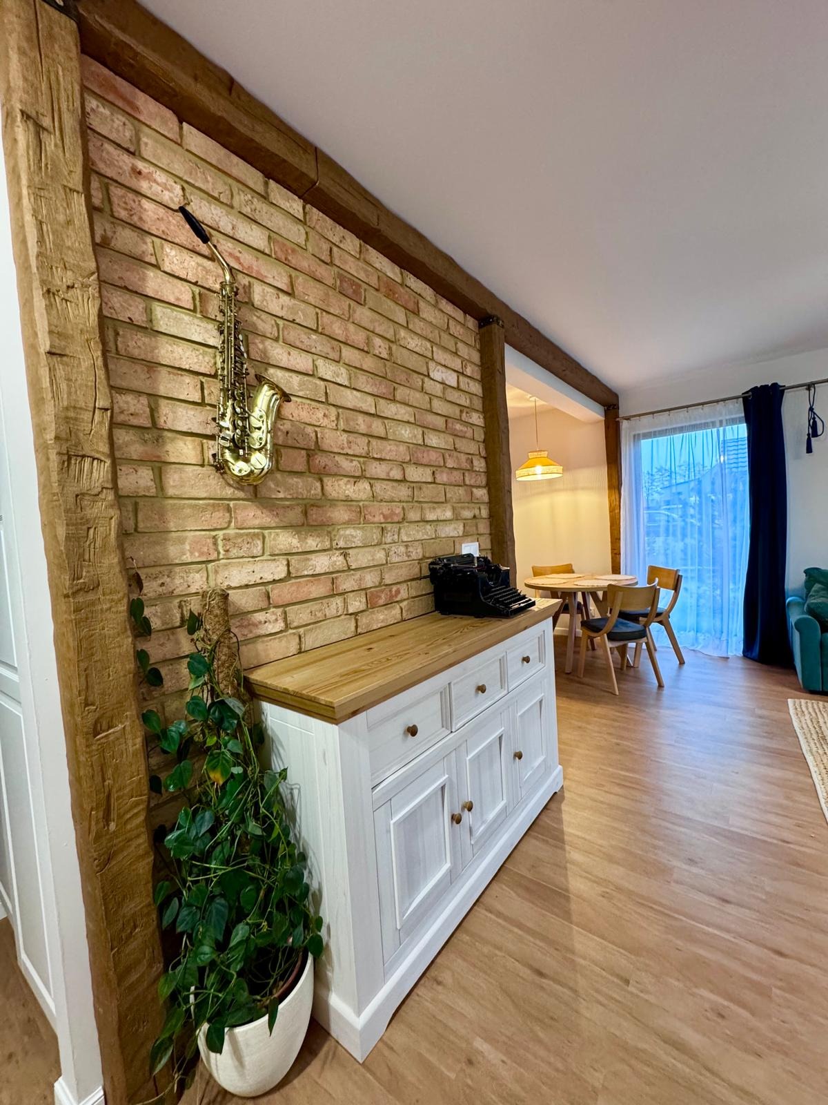 Ceglana ściana z saksofonem i drewnianymi belkami w Mazańcowicach. Biała komoda z drewnianym blatem, roślina doniczkowa i maszyna do pisania tworzą unikalny wystrój.