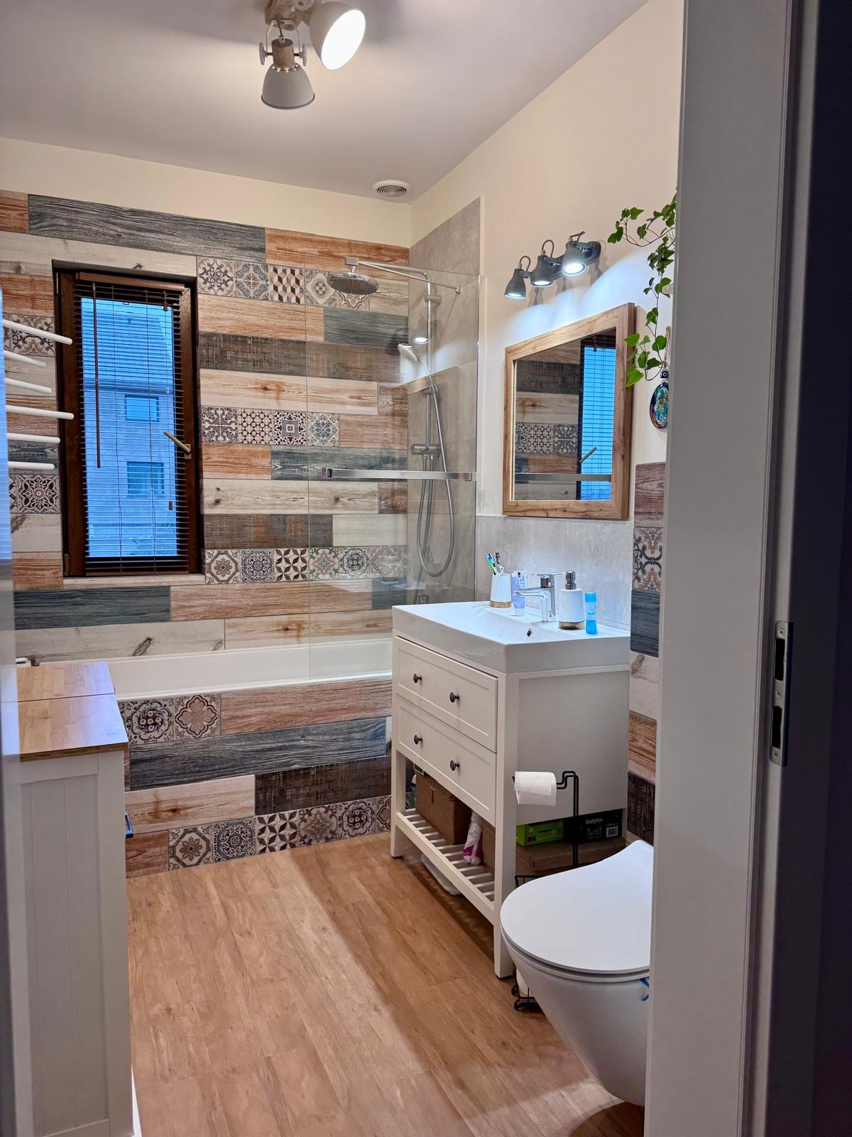 Ukończona łazienka z patchworkowymi płytkami ściennymi w odcieniach brązu i szarości, drewnopodobną podłogą i białą armaturą. Widok zza uchylonych drzwi.