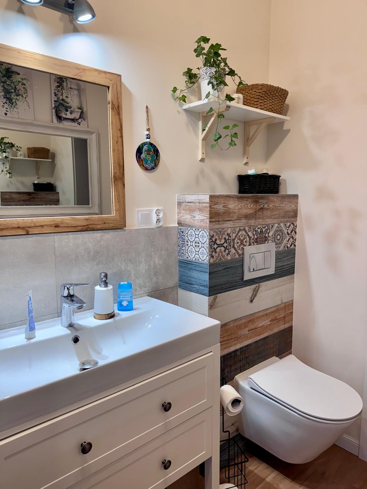 Nowoczesna łazienka z białą szafką pod umywalką, lustrem w drewnianej ramie i toaletą z obudową w patchworkowe wzory. Rośliny doniczkowe dodają świeżości.