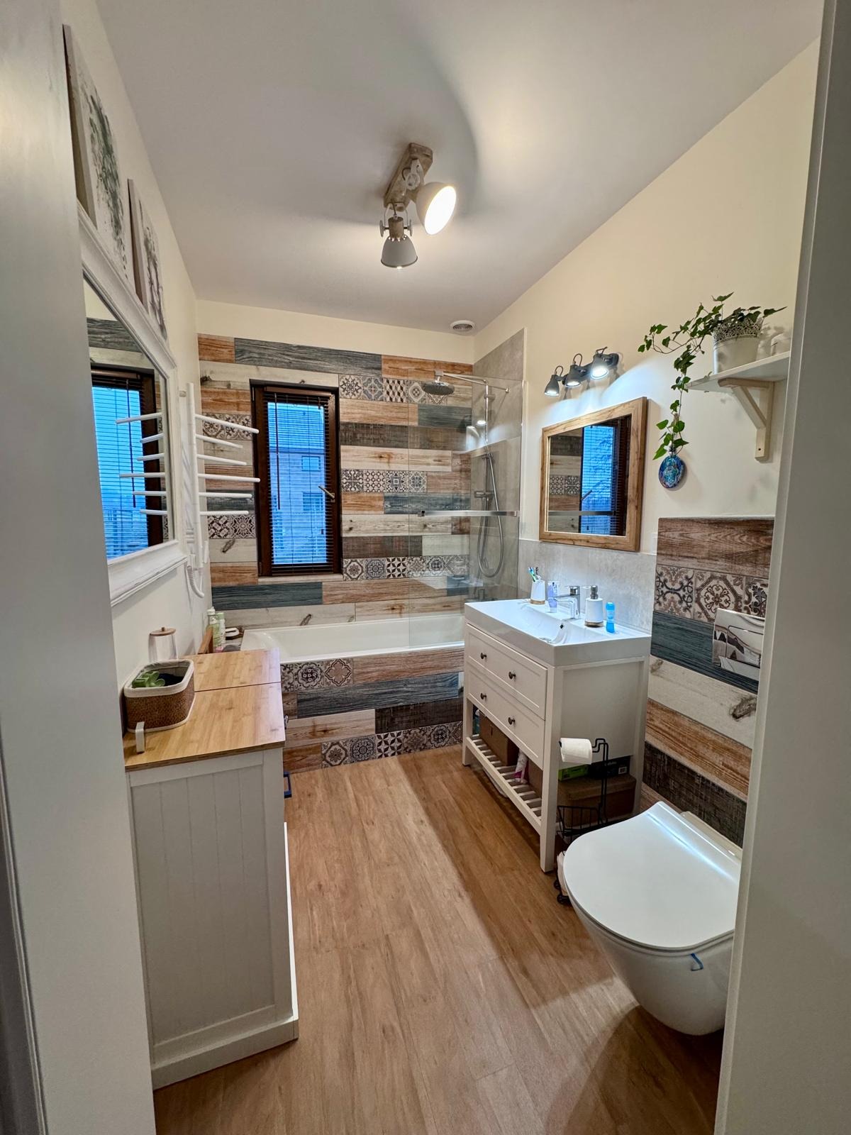 Ukończona łazienka z wanną obudowaną mozaiką, drewnopodobną podłogą i białą szafką umywalkową. Na ścianach dekoracyjne płytki w odcieniach brązu i szarości.