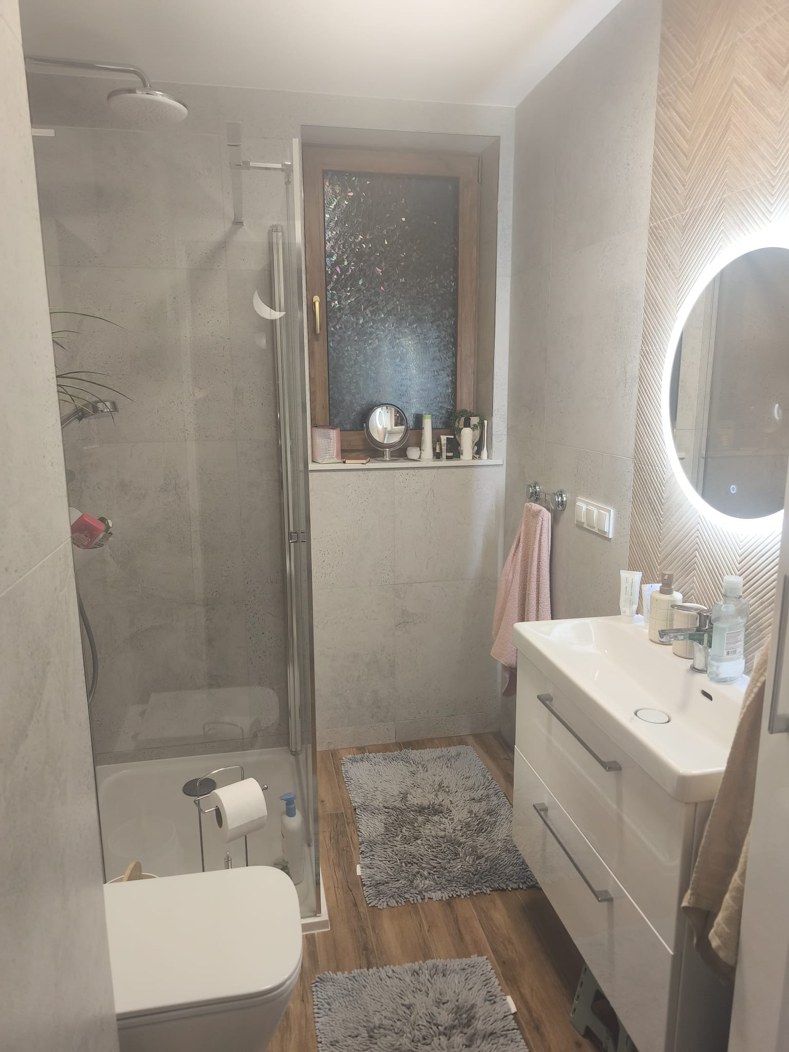 Nowoczesna łazienka z kabiną prysznicową, umywalką z szafką i okrągłym lustrem z podświetleniem LED. Na ścianach szare płytki imitujące beton i drewnopodobna podłoga.