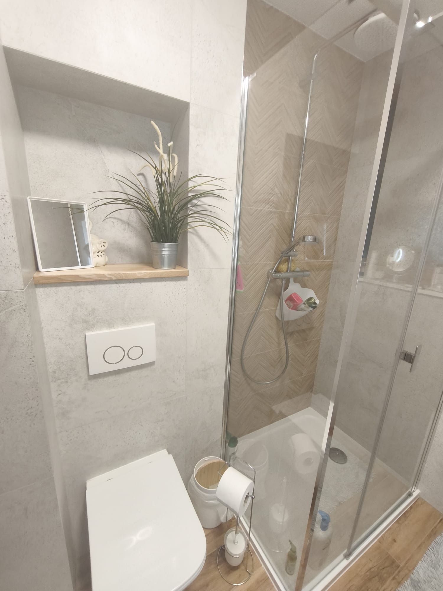 Jasna łazienka po remoncie: kabina prysznicowa ze szklanymi drzwiami, toaleta z podtynkową spłuczką, półka z lustrem i rośliną w metalowej doniczce.