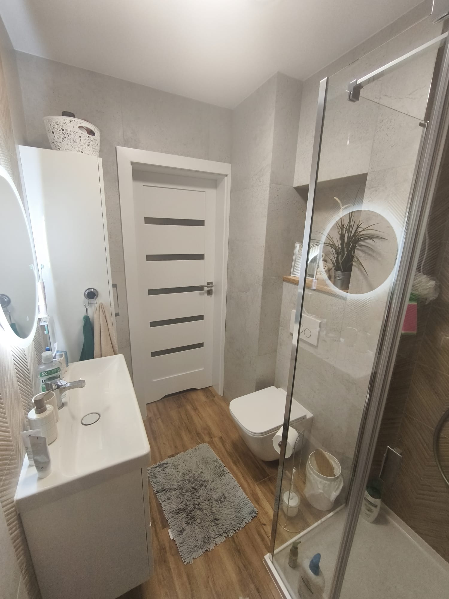 Nowoczesna łazienka z drewnopodobną podłogą, szarą glazurą i białymi meblami. Uwagę przykuwa okrągłe lustro z podświetleniem LED i kabina prysznicowa ze szklanymi drzwiami.