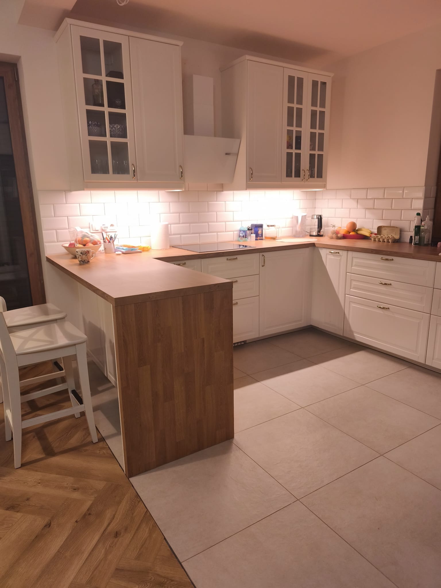 Jasna kuchnia z białymi szafkami, drewnianym blatem i barkiem. Uwagę przykuwają płytki imitujące cegłę nad blatem i kontrastująca podłoga z płytek i parkietu.