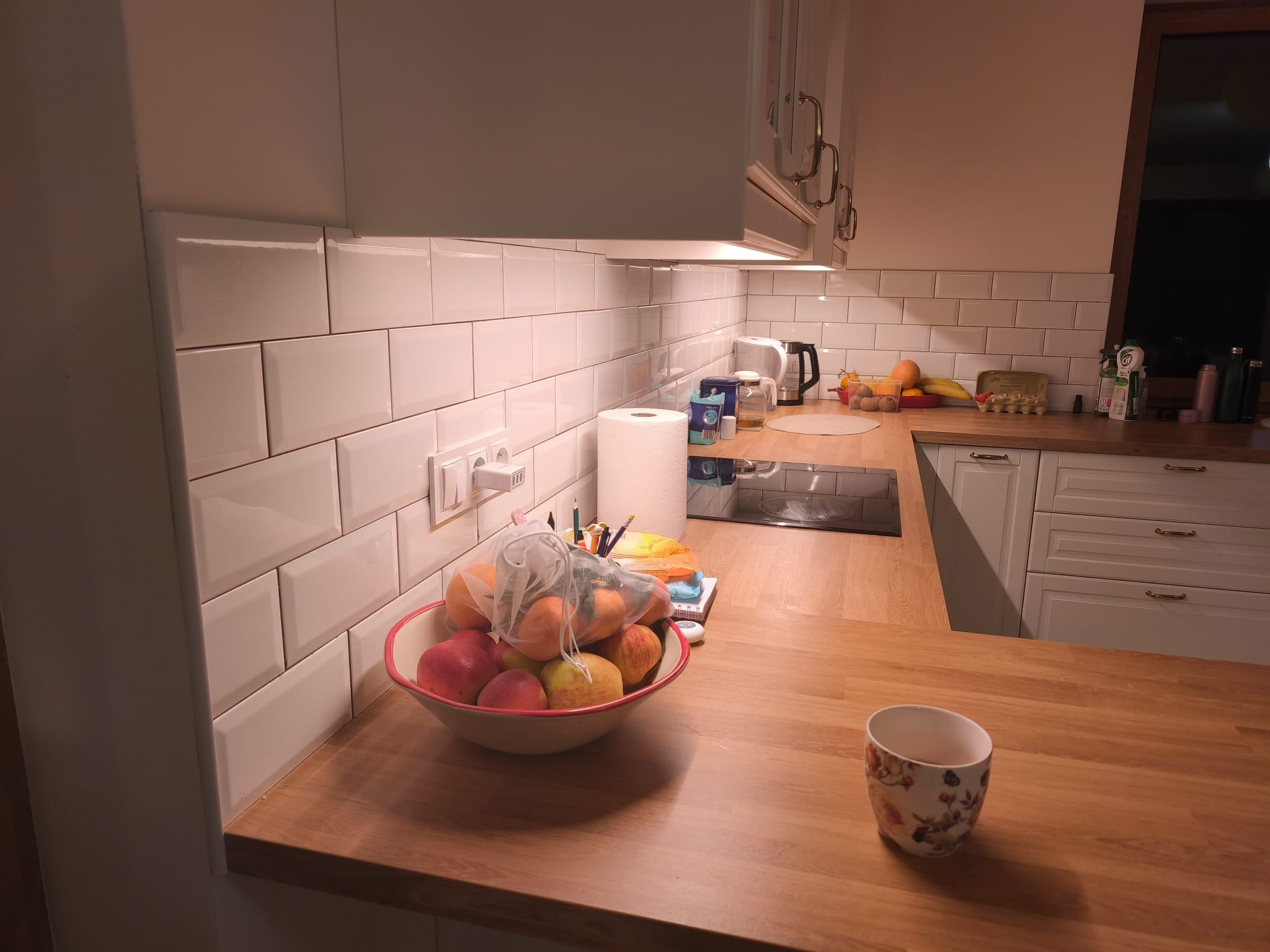 Jasna kuchnia z białymi, prostokątnymi płytkami na ścianie, drewnianym blatem i białymi szafkami. Na blacie misa z owocami i kubek.