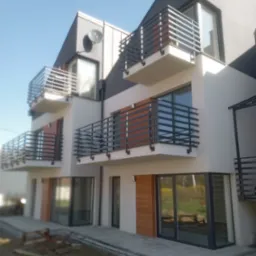 Nowoczesny budynek wielorodzinny z balkonami o grafitowych balustradach. Jasna elewacja z drewnianymi akcentami. Widok z dołu na błękitne niebo.