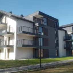 Nowoczesny, trzypiętrowy budynek apartamentowy z balkonami, w odcieniach bieli i szarości, z elementami drewnianymi. Ujęcie z poziomu trawnika w słoneczny dzień.