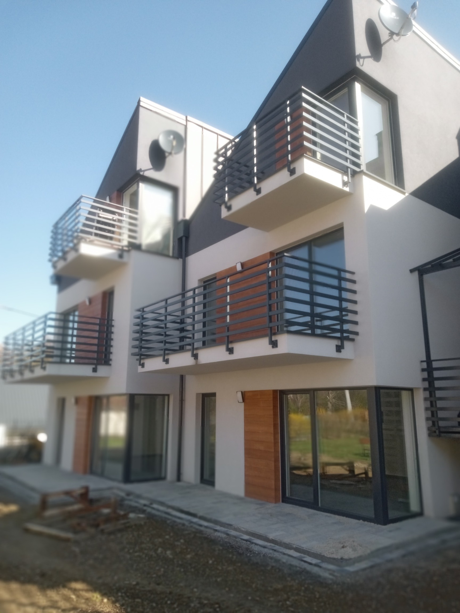 Nowoczesny budynek wielorodzinny z balkonami o grafitowych balustradach. Jasna elewacja z drewnianymi akcentami. Widok z dołu na błękitne niebo.