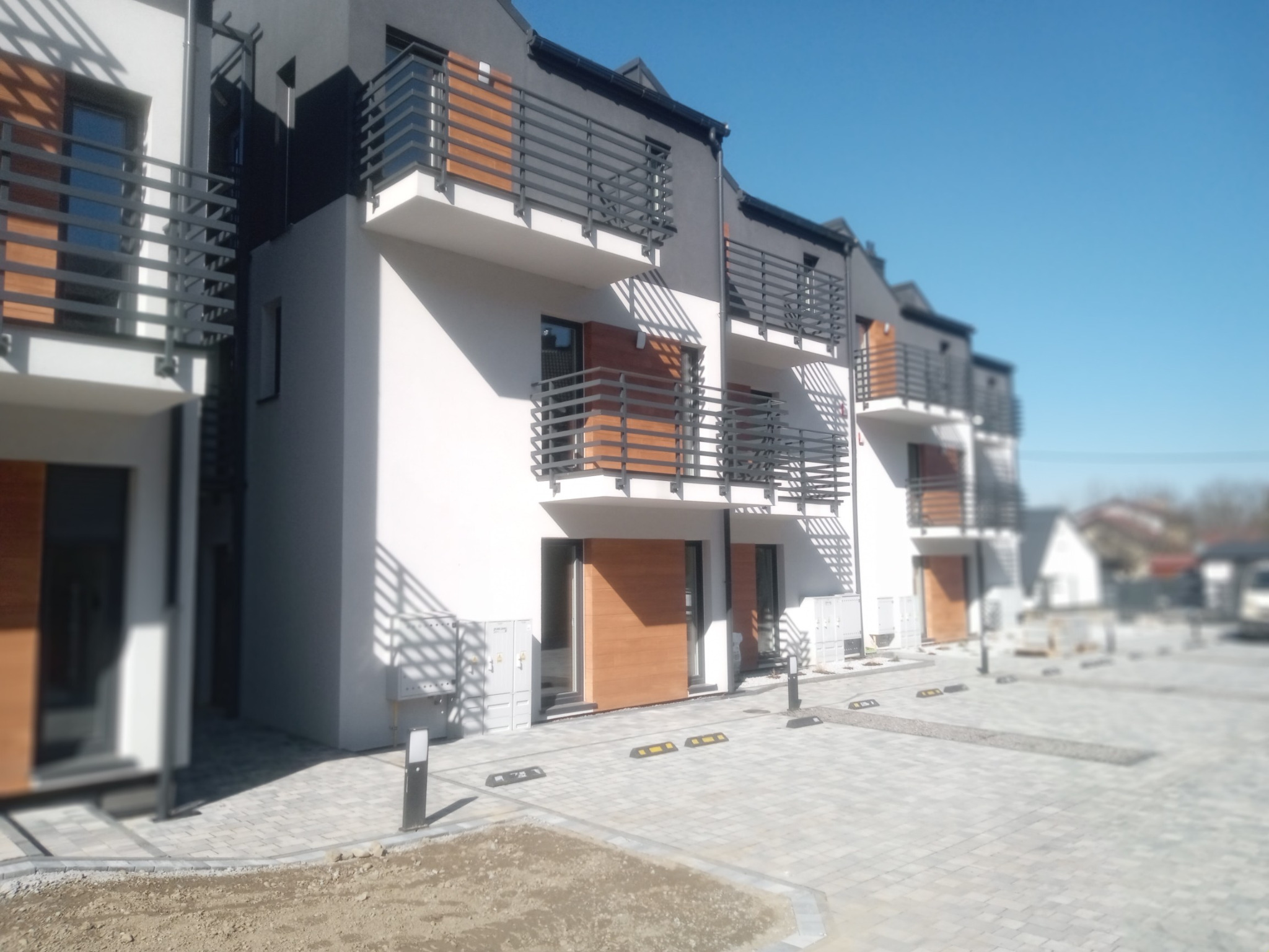 Nowoczesny, szeregowy dom z balkonami i drewnianymi akcentami elewacji. Kostka brukowa na podjeździe, minimalistyczne lampy. Błękitne niebo w tle.