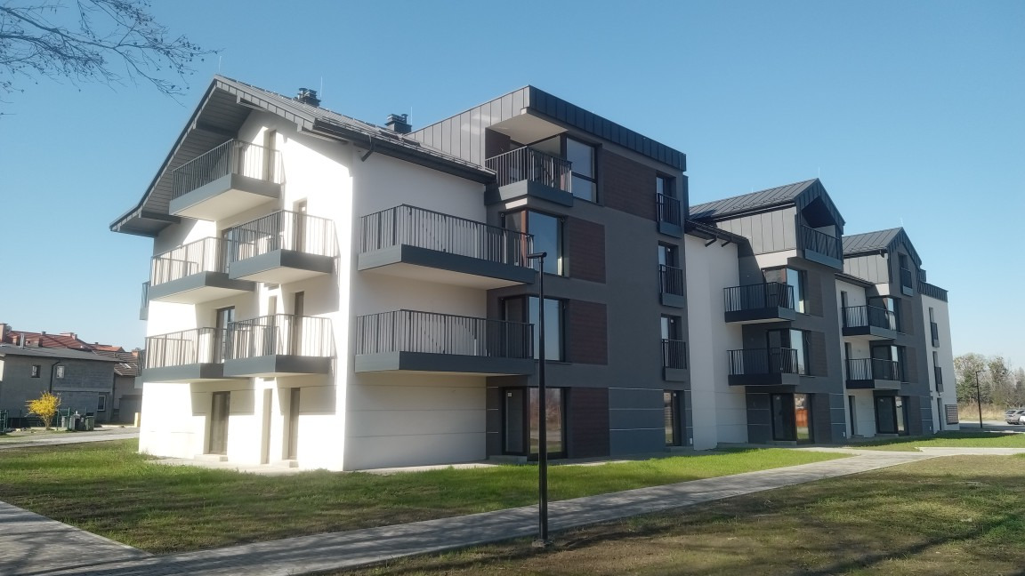 Nowoczesny, trzypiętrowy budynek apartamentowy z balkonami, w odcieniach bieli i szarości, z elementami drewnianymi. Ujęcie z poziomu trawnika w słoneczny dzień.