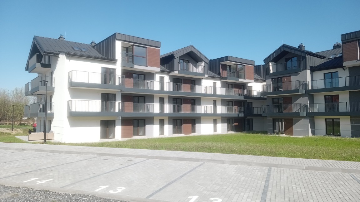 Nowoczesny, trzypiętrowy apartamentowiec z balkonami, z jasną elewacją i ciemnymi elementami wykończenia, widok z parkingu w słoneczny dzień.
