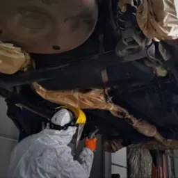 Pracownik w kombinezonie ochronnym, z maską i rękawicami, piaskuje podwozie samochodu na podnośniku. Elementy auta zabezpieczone papierem i taśmą.